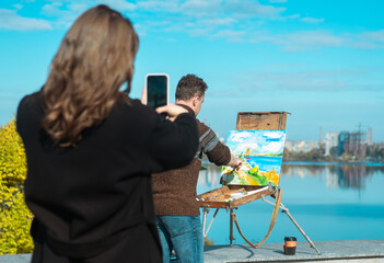 A woman takes a picture of an artist painting a colorful landscape by a lake on a sunny day.