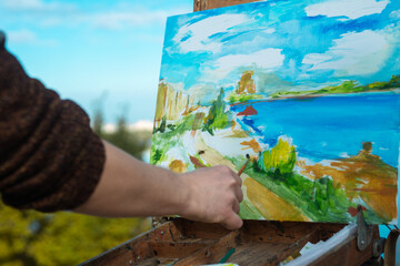 A person painting a colorful seaside landscape on a canvas outdoors. The scene captures nature's beauty.