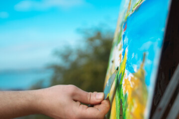 A close-up of an artist using a palette knife to paint a colorful landscape on an easel in an outdoor setting.