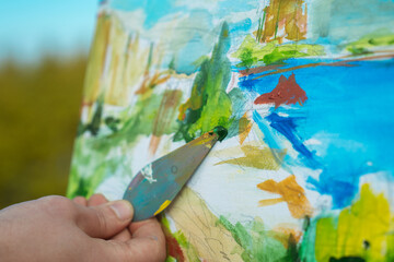 A close-up shot of an artist's hand using a palette knife to create a vibrant painting on canvas outdoors.