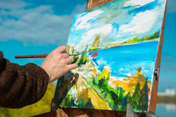 A person painting a colorful seaside landscape on a canvas outdoors. The scene captures nature's beauty.