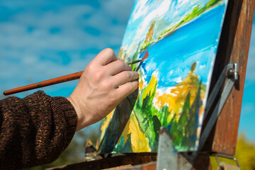 A person painting a colorful seaside landscape on a canvas outdoors. The scene captures nature's beauty.