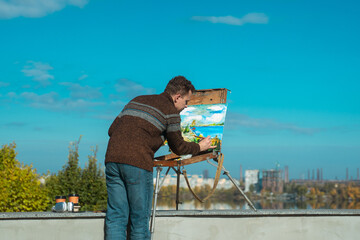 A man paints a vibrant landscape on an easel with a bright blue sky in the background, outdoors.