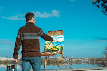 A man paints a vibrant landscape on an easel with a bright blue sky in the background, outdoors.