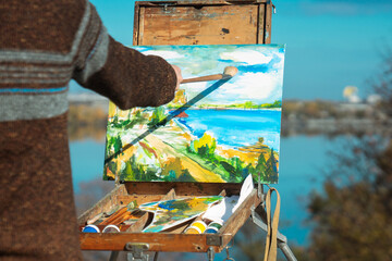A man paints a vibrant landscape on an easel with a bright blue sky in the background, outdoors.