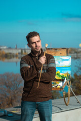 A thoughtful artist stands with a paintbrush next to an easel displaying a landscape painting under a blue sky.