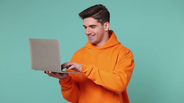 Smart fun young IT man he wears orange hoodie hold in hand use work typing point finger on on laptop pc computer isolated on plain pastel light blue cyan background studio portrait. Lifestyle concept