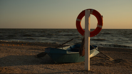 An orange life preserver hangs from a wooden pole next to a turquoise rowboat on a pebble beach....