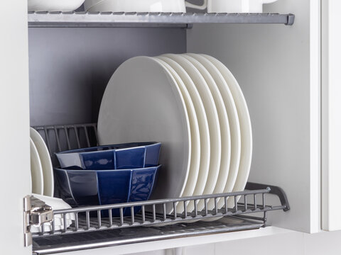 A sleek metal dish-drying rack is neatly integrated into a wall cabinet in a modern kitchen. The rack holds white ceramic kitchenware, plates and mugs, all arranged orderly