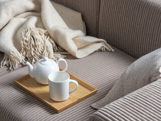 Cozy tea scene with white ceramic teapot and mug on a bamboo tray, resting on a neutral sofa with a soft fringed throw. Hygge-style decor with warm, minimalist textures