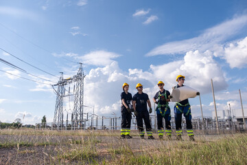 Workers discuss plans at an electrical substation during daytime near power lines and transformer equipment