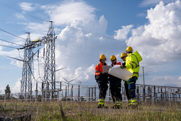 Workers discuss plans at a power station with transmission lines in the background during the day