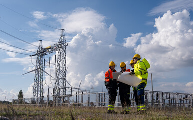 Workers discuss plans near power lines and wind turbines on a clear day in an energy facility
