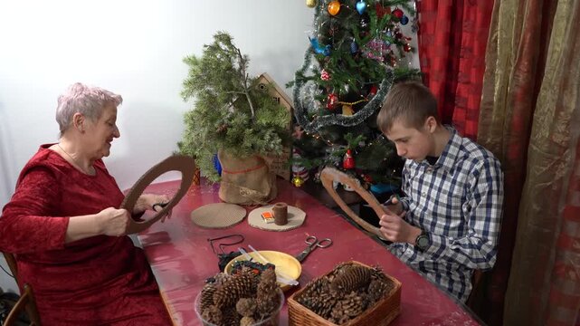 Use scissors to cut out the cardboard circles for the Christmas wreath. Grandmother and grandson make winter craft together. Elderly woman and teenage boy spend time together. Family