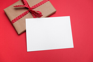 Fototapeta premium Blank paper greeting or invitation card mockup with copy space with gift box featuring a red ribbon on a vibrant red background for festive occasions