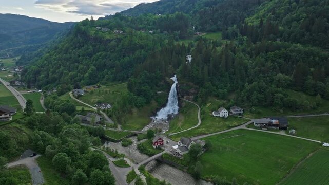 Wide aerial view of Steinsdalsfossen waterfall near Norheimsund, Norway. Drone footage capturing rural landscape, farmland, forested hills, and flowing water in summer.
