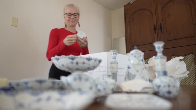 Mature woman unpacks her inherited china dining sets while recalling memories of family dinners and the value of treasured items.