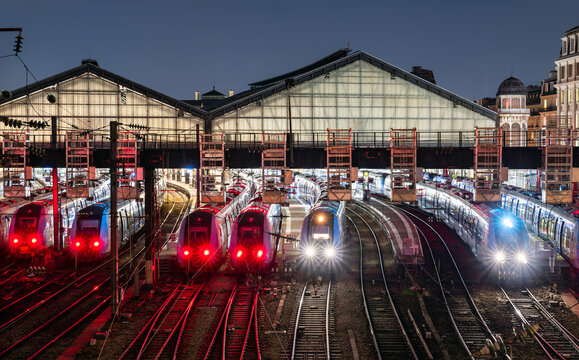 Gare Saint-Lazare station in Paris, France. Night view features railway tracks and illuminated commuter trains at the platforms under the historic glass roof