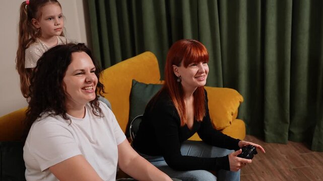 Two women and young girl are having fun playing, enthusiastically competing in video games in comfort of own home. Family playing on game console with joystick or controller, spending weekend together