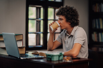 Student drinking coffee while studying in library using laptop