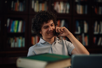 Student talking on smartphone and smiling in library with books and laptop