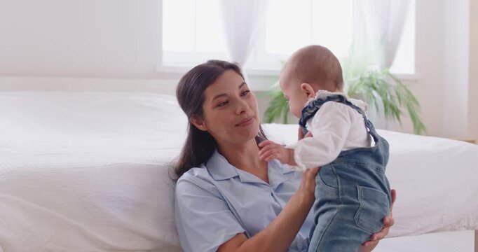 Happy family time, help and support from mother to cute baby in first steps at home. Young woman sitting by bed, helping child stand up and speaking gentle words, standing boy hugging mom with trust