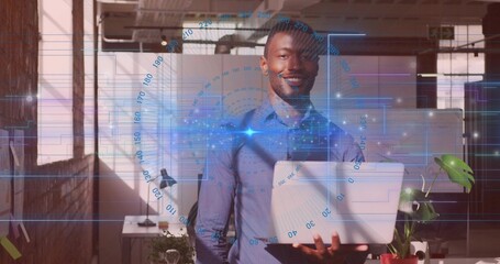 Smiling man in light blue shirt holding laptop in open-plan office, showing holographic overlay