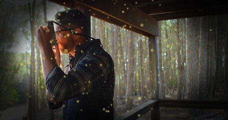 Scanning bearded man holding binoculars on wooden porch by railing in cap and vest, golden motes