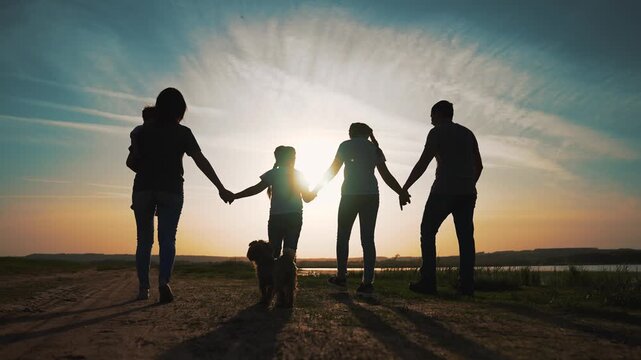 Family walking hand in hand at sunset on dirt road by lake, silhouette of parent and child with dog, holding hand casting long shadow across grass under dramatic sky and golden reflective water