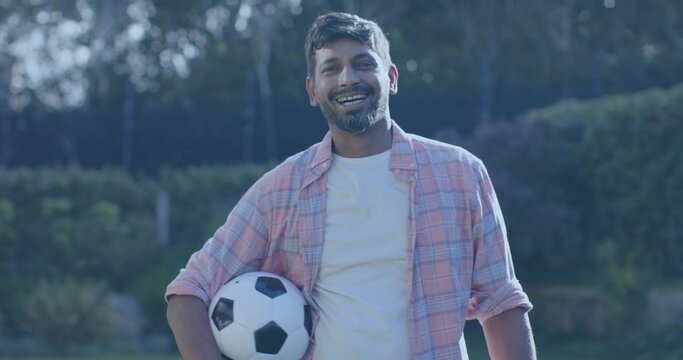 Smiling man wearing light pink plaid shirt, white tee, holding black-and-white soccer ball on lawn - Powered by Adobe