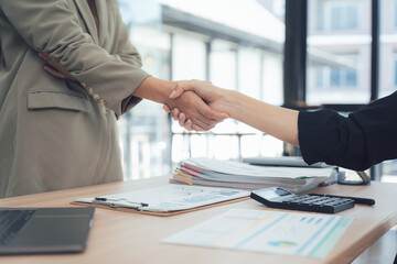 Handshake Deal: A close-up shot of two individuals engaged in a handshake, sealed a business agreement, conveying trust and partnership. They stand at a wooden desk with office tools.