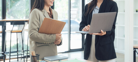 Business Colleagues in Collaboration: Two professional women engage in a dynamic business discussion within a modern office setting. One woman is holding a document folder.