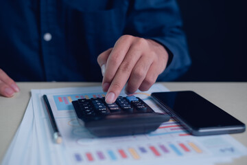 Business finance concept with close up of hand using calculator on financial report documents next to smartphone on desk, analyzing budget, accounting data, investment planning,