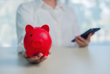Red piggy bank held in hand with smartphone in background, representing digital saving, personal finance, smart budgeting, mobile banking, financial technology, money management, modern lifestyle.