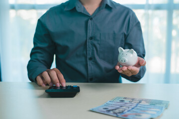 Person using calculator while holding white piggy bank with cash on desk, symbolizing saving money, budgeting, personal finance, expense control, financial planning, investment strategy, and security.