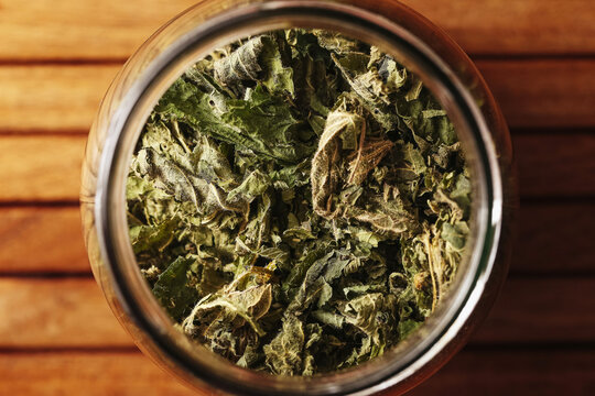 Dried nettle leaves in glass jar viewed from above on wooden table. Herbal tea ingredient for infusion preparation. Medicinal plant storage closeup. Natural herbal remedy concept.