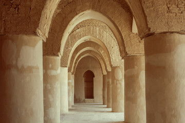 A series of ancient, earth-toned stone arches supported by thick pillars create a rhythmic perspective down a long corridor in a historic structure