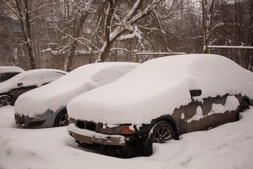 Multiple cars completely buried under thick snow in a parking lot after a heavy winter storm, with windshield wipers raised to prevent freezing.