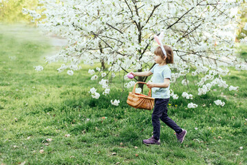 Easter Egg Hunt with Child Carrying Bright Plastic Eggs. Smiling child wearing bunny ears holding a wicker basket filled with colorful Easter eggs in a blooming spring garden