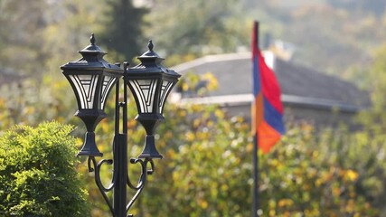 A vintage black lamppost stands in a sunny park. In the blurred background, the Armenian flag waves gracefully amidst lush green and yellow autumn trees and foliage.