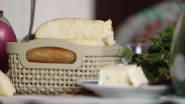 Close-up view of a plastic basket filled with fresh white bread slices on a table, accompanied by cheese cubes and green herbs in the soft-focused background during meal.

