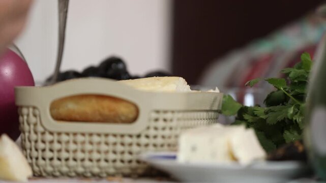 Close-up view of a plastic basket filled with fresh white bread slices on a table, accompanied by cheese cubes and green herbs in the soft-focused background during meal.

