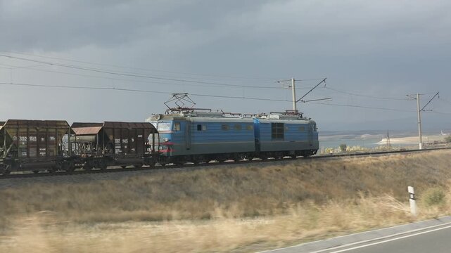 Freight train consisting of brown hopper wagons and a blue electric locomotive moves along the railway track against a cloudy sky background in a rural landscape.

