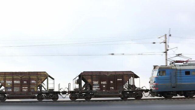 Freight train consisting of brown hopper wagons and a blue electric locomotive moves along the railway track against a cloudy sky background in a rural landscape.

