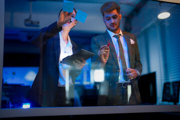 A businessman writing ideas on a glass wall with colorful markers. The woman holds a digital tablet while the man writes Marketing Concept in red.