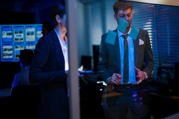 Innovative digital transformation team brainstorming on glass wall. Professional executive writing future business strategy while female colleague analyzes data in modern blue-lit office at night.