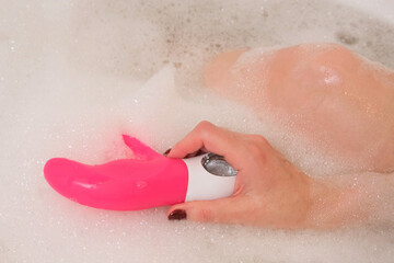 Female hand lowers pink sex toy onto soap suds filling tub