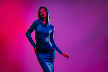 Young stylish woman in a blue dress with a hood posing in neon lights on a colorful pink and purple...