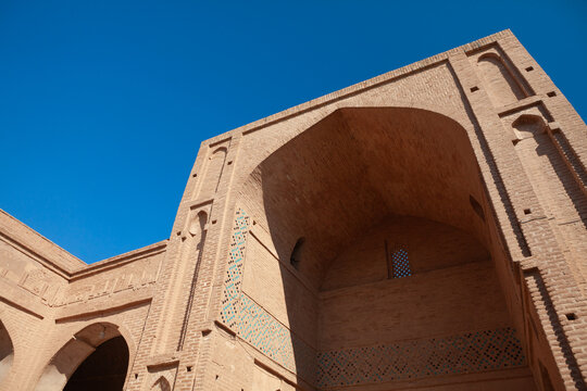 A low-angle shot of a majestic, earth-toned brick iwan with intricate geometric tilework under a clear blue sky, showcasing the grand scale of Islamic architecture.