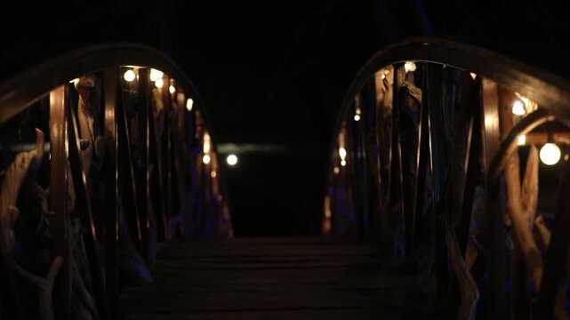 Atmospheric view of a rustic wooden footbridge with branch railings illuminated by string lights at night. The warm glow creates a romantic and magical path through darkness.

